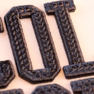 May include: Close-up of black embroidered letters spelling "LOL". The letters have a textured appearance and are stitched onto a light-colored fabric. The embroidery has a detailed, woven pattern.