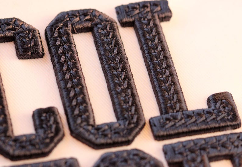 May include: Close-up of black embroidered letters spelling "LOL". The letters have a textured appearance and are stitched onto a light-colored fabric. The embroidery has a detailed, woven pattern.