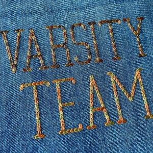 May include: Embroidered denim patch with the text "Varsity Team" in brown, orange, and green thread.
