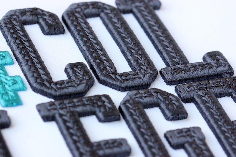 May include: Close-up of embroidered letters spelling out "GOT IT" in a dark gray color. The letters have a textured, woven appearance, and are stitched onto a white fabric. A few teal letters are also visible.