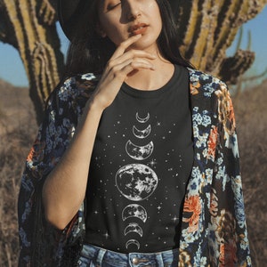 Puede incluir: Camiseta negra con un dise&ntilde;o blanco de las fases lunares y estrellas. La persona lleva un sombrero negro y un kimono con estampado floral. El fondo muestra un cactus y un paisaje des&eacute;rtico.