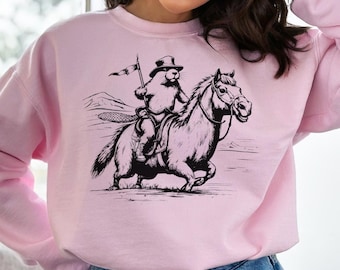 Peut inclure: Un sweat-shirt rose avec une illustration en noir et blanc d'un castor portant un chapeau haut de forme et monté sur un cheval. Le castor tient un drapeau.