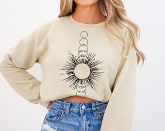 Peut inclure: Un sweat-shirt court beige avec un motif soleil et lune noir et blanc. Le motif représente un soleil avec des rayons et une ligne verticale de lunes.