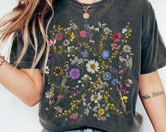 Może przedstawiać: Szary t-shirt z nadrukiem kwiatowym, przedstawiającym różne suszone kwiaty w odcieniach żółtego, różowego, fioletowego i niebieskiego. Koszulka ma okrągły dekolt i krótkie rękawy.