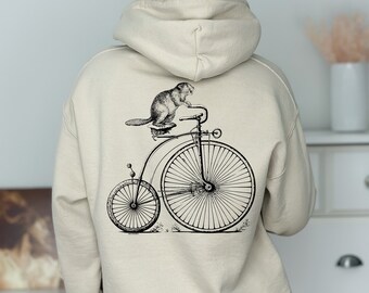 Peut inclure: Un sweat à capuche beige avec une illustration en noir et blanc d'un castor à vélo vintage sur le dos.