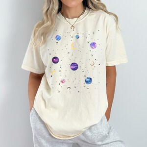 May include: A cream-colored t-shirt with a graphic print of planets and stars in shades of blue, purple, and pink.