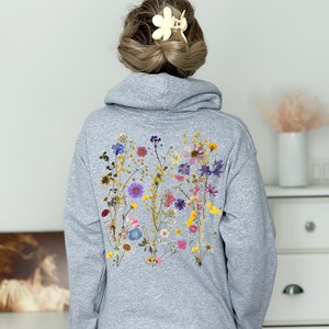 May include: A grey hoodie with a floral design on the back. The design features a variety of pressed flowers in shades of pink, purple, yellow, and white.