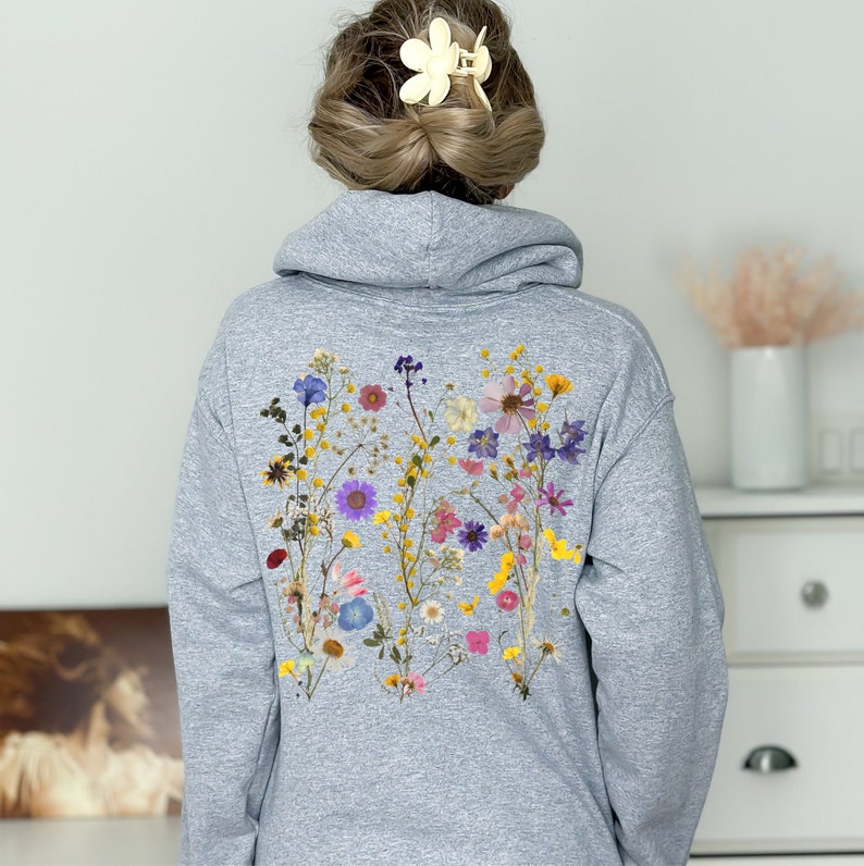 May include: A grey hoodie with a floral design on the back. The design features a variety of pressed flowers in shades of pink, purple, yellow, and white.