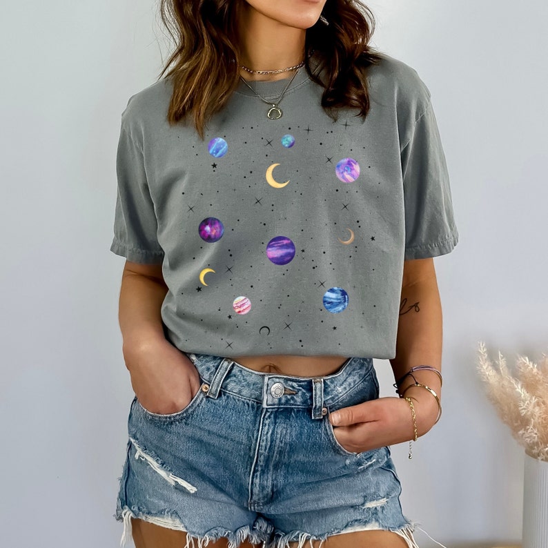 May include: A gray t-shirt with a galaxy print featuring planets, stars, and moons. The shirt is cropped and has a relaxed fit.