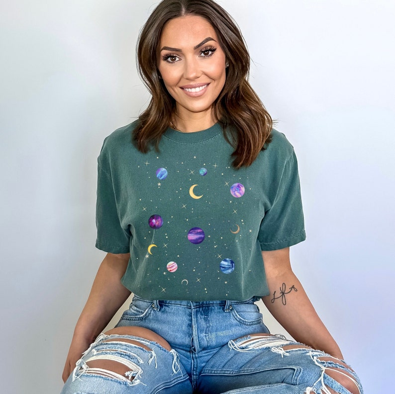 May include: A woman wearing a green t-shirt with a celestial print featuring planets, stars, and a crescent moon. The shirt has a relaxed fit and short sleeves.