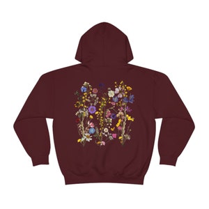 May include: Burgundy hooded sweatshirt with a floral print on the back. The print features a variety of dried flowers in shades of pink, purple, yellow, and blue.