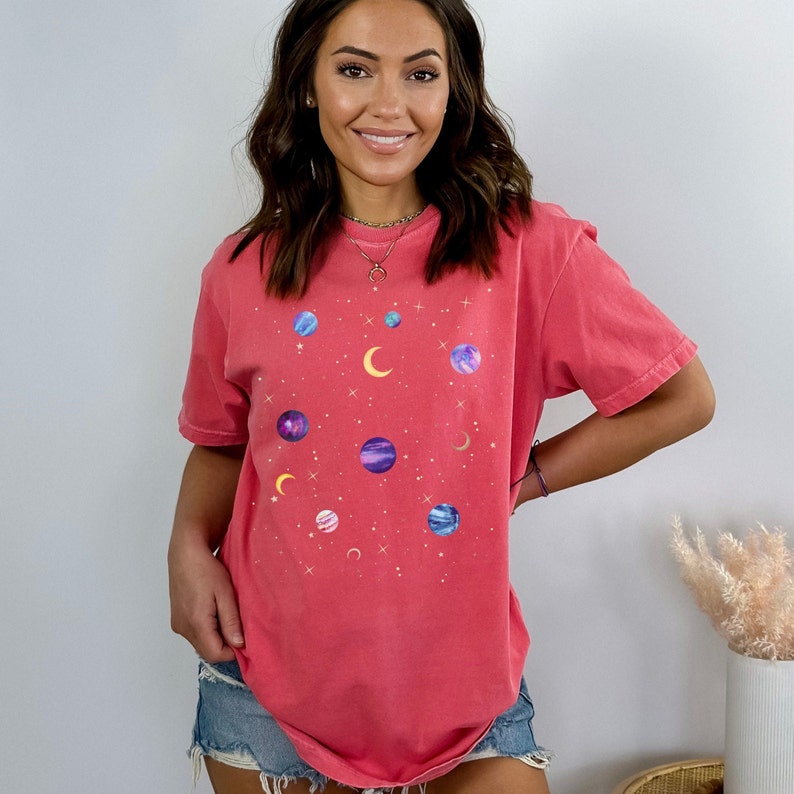 May include: A coral pink t-shirt with a graphic print of planets and stars. The shirt has a relaxed fit and a crew neck.