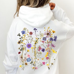May include: White hoodie with a floral design on the back. The design features a variety of pressed flowers in shades of pink, purple, yellow, and white.
