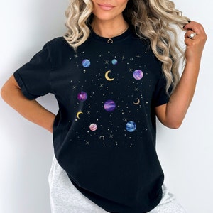 May include: Black t-shirt with a galaxy print featuring multiple planets and moons in shades of purple, blue, and pink. The planets are surrounded by gold stars and crescent moons.