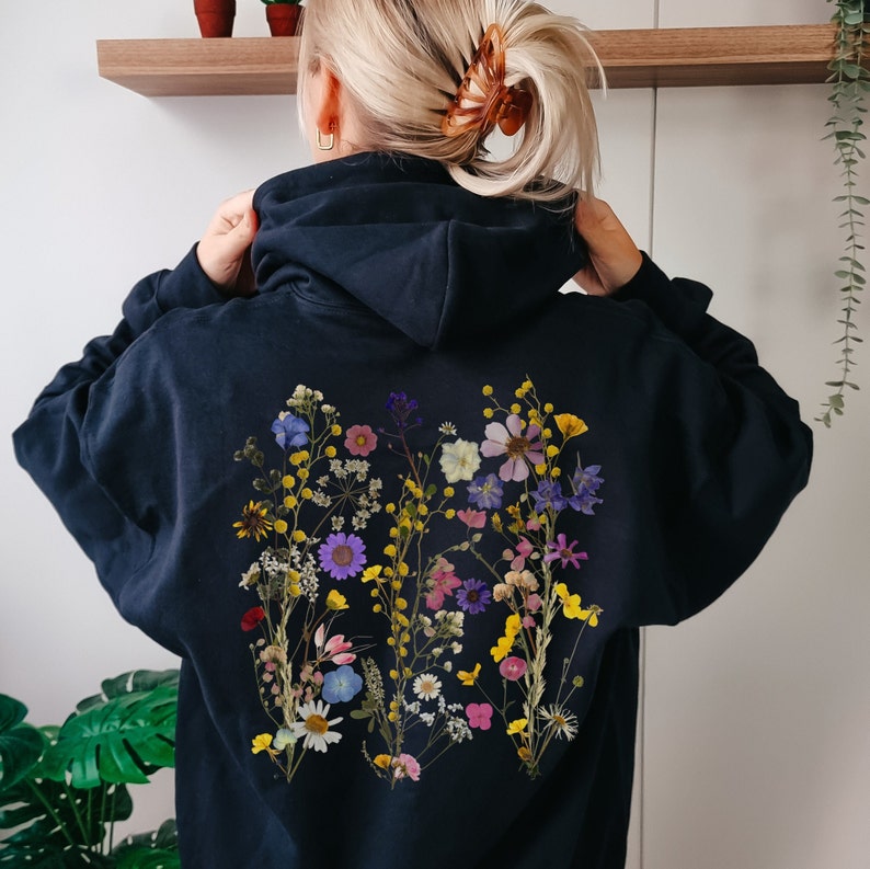 May include: A black hoodie with a floral design on the back. The design features a variety of pressed flowers in shades of pink, purple, yellow, and white.