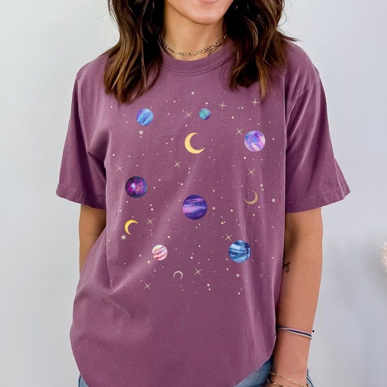 May include: A purple t-shirt with a galaxy print featuring planets, stars, and moons. The shirt has a relaxed fit and short sleeves.