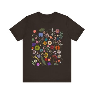 Może przedstawiać: Ciemnobrązowy t-shirt z kolorowym nadrukiem kwiatowym. Nadruk przedstawia r&oacute;żne kwiaty, liście i grzyby w odcieniach r&oacute;żowego, fioletowego, niebieskiego, ż&oacute;łtego, pomarańczowego i czerwonego.