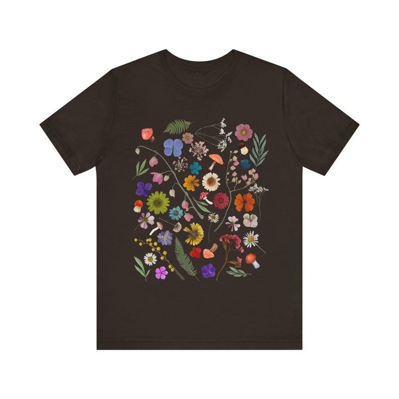 Może przedstawiać: Ciemnobrązowy t-shirt z kolorowym nadrukiem kwiatowym. Nadruk przedstawia r&oacute;żne kwiaty, liście i grzyby w odcieniach r&oacute;żowego, fioletowego, niebieskiego, ż&oacute;łtego, pomarańczowego i czerwonego.