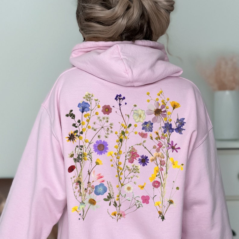 May include: Pink hoodie with a floral design on the back. The design features a variety of pressed flowers in shades of pink, purple, yellow, and blue.