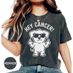 May include: A charcoal gray short-sleeve t-shirt featuring a white graphic of a dog wearing sunglasses and giving the middle finger with both hands. The text above the dog reads HEY CANCER! in white capital letters. The shirt has a vintage wash.