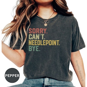 May include: A dark gray cropped t-shirt with the text "SORRY. CAN'T. NEEDLEPOINT. BYE." in a vintage style font with colorful accents.