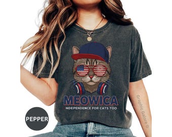 May include: Dark gray t-shirt featuring a cat wearing sunglasses with an American flag design, a blue and red baseball cap, and headphones. The shirt has the text "MEOWICA" and "INDEPENDENCE FOR CATS TOO".