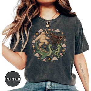 May include: A charcoal gray t-shirt featuring a circular graphic print of a white yeti, a brown moth creature, a green sea monster, and a green alien dancing together. The design includes small pink flowers, clouds, and yellow stars on a dark background.