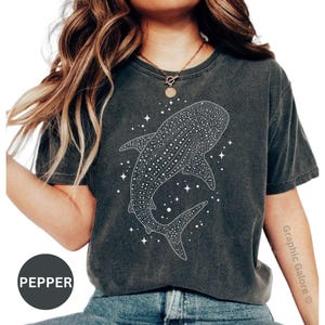 May include: Dark gray t-shirt featuring a detailed white illustration of a whale shark with a starry background. The shirt has a crew neck and short sleeves. The word "PEPPER" is printed on a black circle.