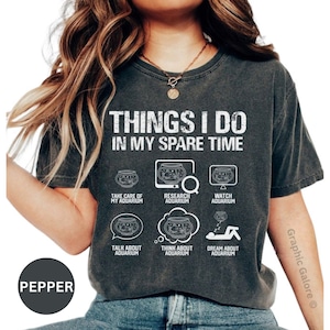 May include: A dark gray t-shirt with the text "THINGS I DO IN MY SPARE TIME" and a series of illustrations of fish tanks with text describing activities related to aquariums. The text includes "TAKE CARE OF MY AQUARIUM", "RESEARCH AQUARIUM", "WATCH AQUARIUM", "TALK ABOUT AQUARIUM", "THINK ABOUT AQUARIUM", and "DREAM ABOUT AQUARIUM".