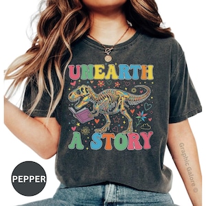 May include: Dark gray t-shirt with the words "UNEARTH A STORY" in colorful letters. A dinosaur skeleton is reading a book, surrounded by hearts and flowers. The shirt is a vintage style.