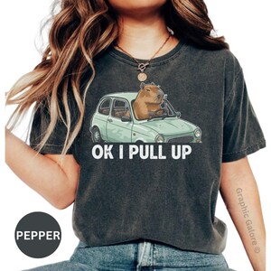 May include: Dark gray t-shirt featuring a capybara driving a light green car. The text "OK I PULL UP" is printed in white. The shirt is a Pepper color.