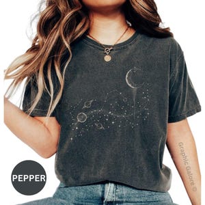 May include: Dark gray t-shirt featuring a celestial design with a crescent moon, planets, and stars. The shirt has a vintage, washed look. The word "PEPPER" is printed on a black circle.
