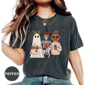 May include: A charcoal gray short-sleeve t-shirt featuring a graphic print of a ghost with a leopard print bow, a skeleton wearing a witch hat, and a pumpkin-headed figure in a sweater. The text PEPPER is visible in a circular badge.