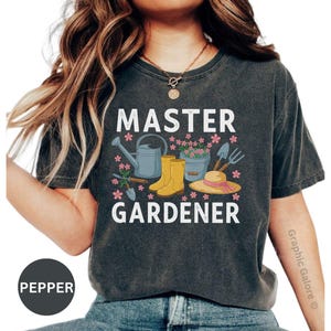 May include: Dark gray t-shirt with the words "MASTER GARDENER" in white, featuring gardening tools and flowers. The design includes a watering can, boots, a hat, and a trowel. The shirt is a comfortable, casual style.