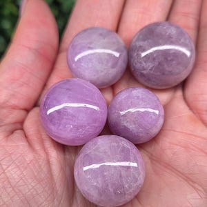 May include: Five polished, spherical gemstones in varying shades of lavender and purple are held in a hand. The stones have a smooth, reflective surface, with subtle variations in colour and texture. The gemstones are a natural product.