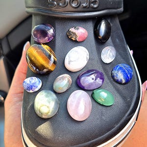 May include: Black Crocs shoe adorned with various colorful gemstones. The gemstones include amethyst, tiger's eye, and rose quartz. The shoe has a white stripe along the sole and the word "CROCS" is embossed on the strap.