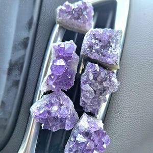 May include: Six amethyst crystal clusters in varying sizes and shapes are arranged on a silver and black car vent. The crystals display a range of purple hues, from light lavender to deep violet, with visible crystalline structures.