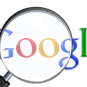 May include: A magnifying glass focuses on the word "Google" in red, yellow, blue, and green, set against a white background. The magnifying glass has a black handle and a clear lens, emphasizing the search engine's logo.