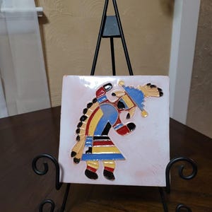 May include: A square ceramic tile featuring a colorful, raised relief depiction of a flute player in blue, yellow, red, and black on a white background, displayed on a black metal stand with scrollwork feet and a tall, pointed support frame.