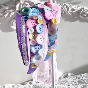 以下が含まれることがあります： 紫色のヘッドバンドに、さまざまな色のボタンが取り付けられています。ボタンはピンク、ブルー、イエロー、ホワイトの色合いです。ヘッドバンドは白いレースのトリムで飾られています。