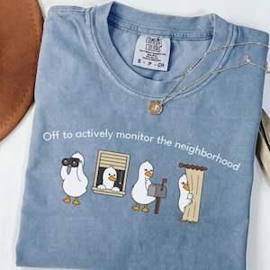 Op de afbeelding: Een lichtblauw Comfort Colors T-shirt met de tekst "Off to actively monitor the neighborhood." Het shirt heeft een cartoon eendenontwerp met een verrekijker, die uit een raam gluurt, bij een brievenbus en achter een gordijn. Een gouden ketting is over het shirt gedrapeerd.