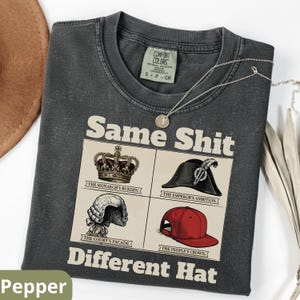 Könnte beinhalten: Dunkelgraues T-Shirt mit dem Aufdruck "Same Shit Different Hat" und Illustrationen einer Krone, eines Zweispitzhutes, einer Perücke und einer Baseballkappe. Das Shirt hat ein Comfort Colors-Etikett und eine silberne Halskette.