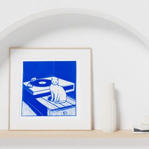 May include: A framed blue and white print of a cat sitting on a record player. The print is on a white shelf with a white vase and a stack of books.