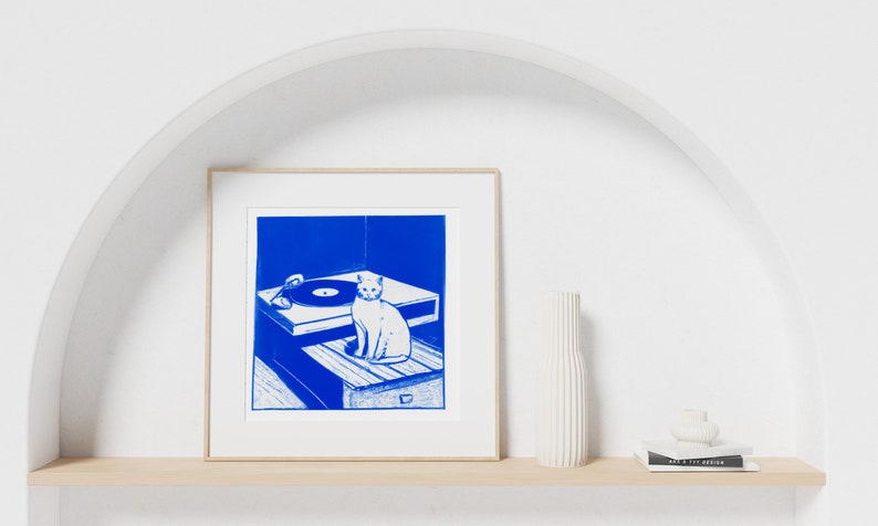 May include: A framed blue and white print of a cat sitting on a record player. The print is on a white shelf with a white vase and a stack of books.