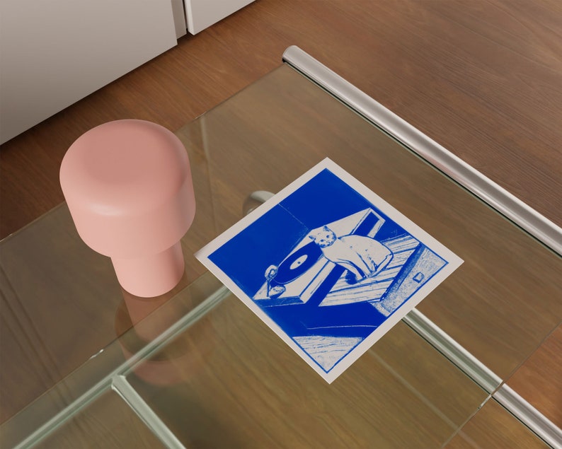 May include: A blue and white illustration of a cat sitting on a record player. The illustration is printed on a white card and is sitting on a glass table.
