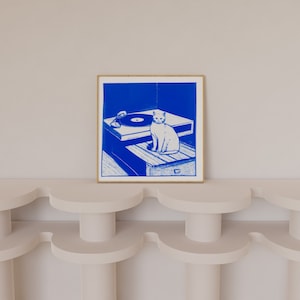 May include: A blue and white illustration of a cat sitting on a record player. The cat is looking to the right. The record player is on a wooden table. The illustration is framed in a light wood frame.