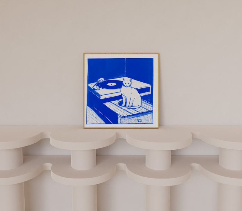 May include: A blue and white illustration of a cat sitting on a record player. The cat is looking to the right. The record player is on a wooden table. The illustration is framed in a light wood frame.