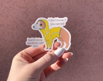 May include: A sticker featuring a cartoon dog wearing a yellow hooded jacket. The dog is tan and white with the text "I don't know if you can tell" and "but I'm being very brave right now". The sticker is held by a hand against a pink background.