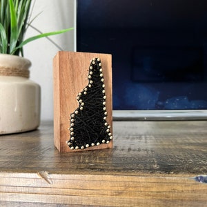 May include: A wooden block with a black string art design of the state of New Hampshire. The string art is outlined with gold tacks.