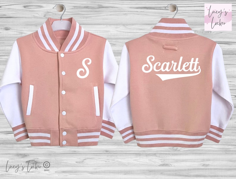 May include: A dusty rose varsity jacket with white sleeves and white striped ribbed trim at the collar, cuffs, and hem. The front features a white letter S, while the back displays the white text Scarlett in a script font with a line underneath.